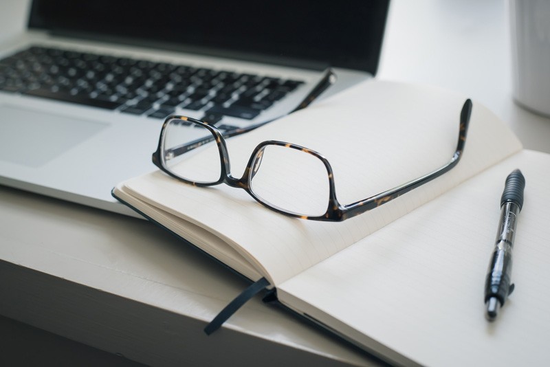 A laptop with an opened blank book with a pair of upturned  glasses and pen resting on the pages.