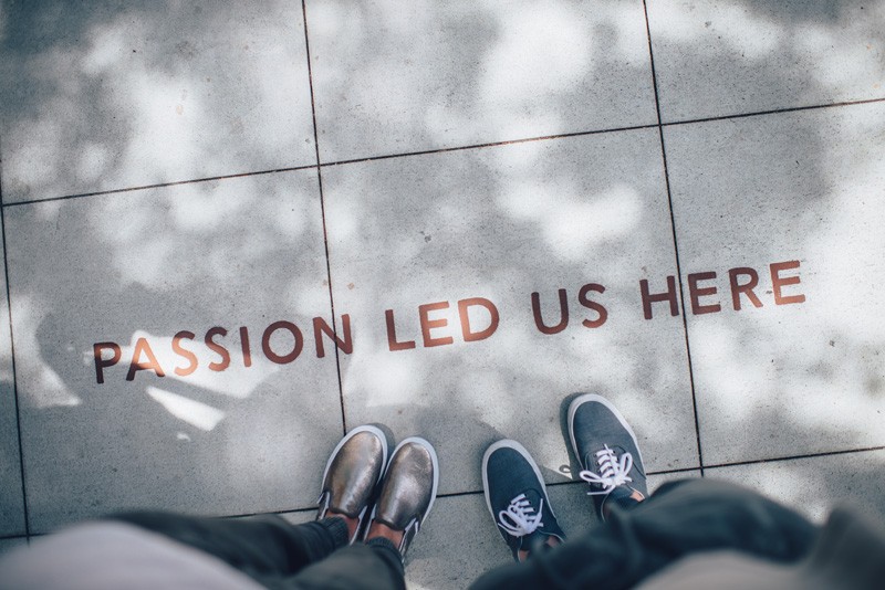 Birds-eye view of two pairs of enclosed feet standing on light grey pavers with the words 'Passion led us here'.