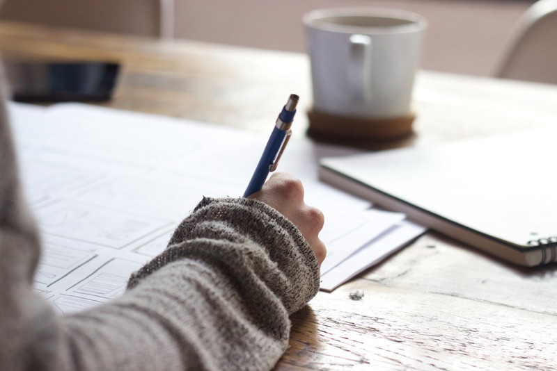 A woman's hand writing on paper on a wooden table. A cup and notebook is lying beside her hand.